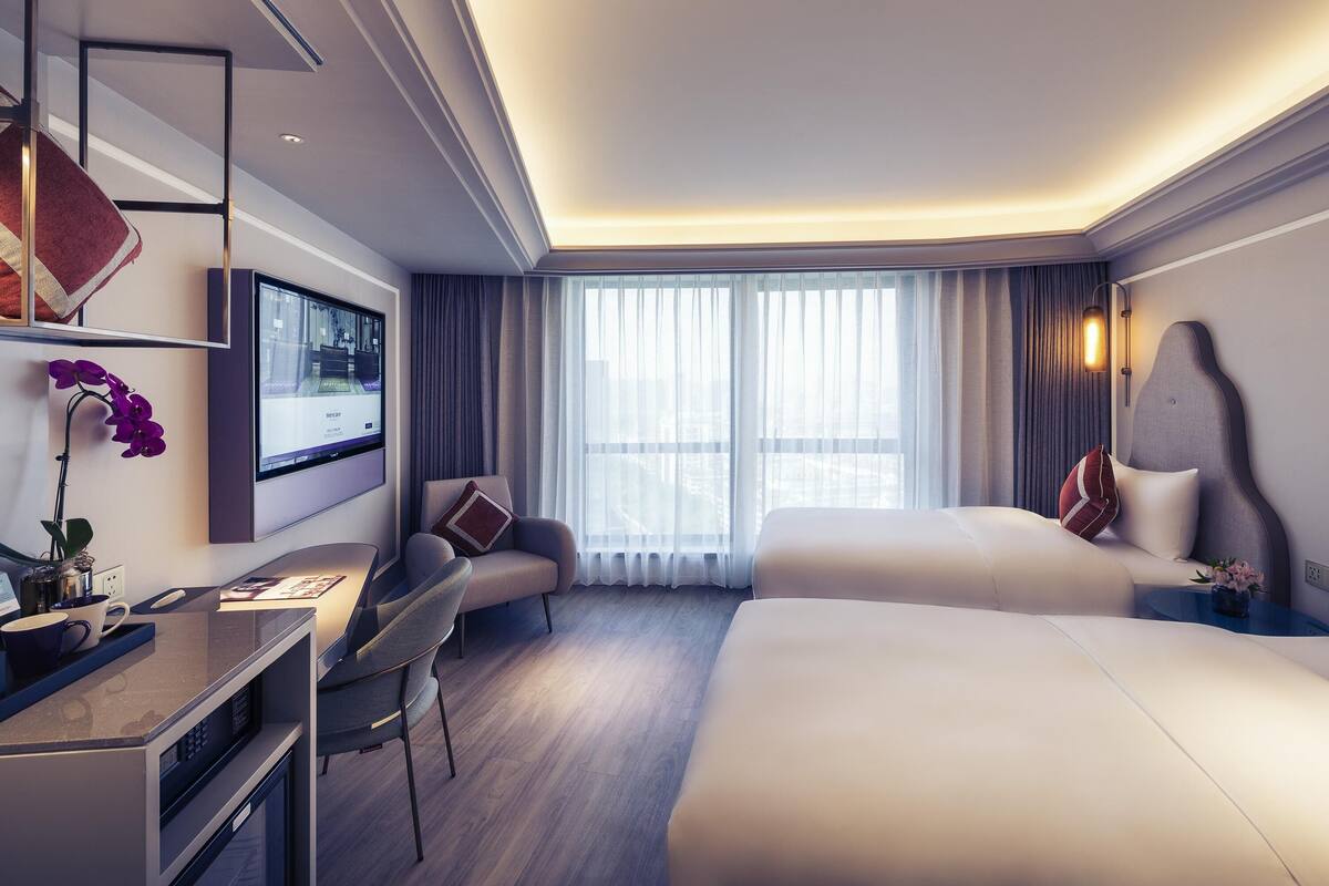 Une chambre d’hôtel moderne équipée d’un téléviseur à écran plat, d’un bureau, de deux lits et d’une grande fenêtre avec des rideaux.