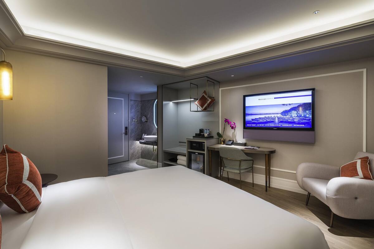 Une chambre d’hôtel moderne, dotée d’un grand lit, d’un bureau, d’une télévision et d’une salle de bain visible à travers une porte ouverte.