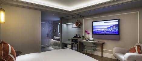 A modern hotel room with a large bed, a desk with a computer, a TV mounted on the wall, and a bathroom visible through an open door.