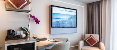 Ett modernt hotellrum med ett skrivbord, en bekväm fåtölj, en digital klocka, en kaffemaskin och ett stort fönster med gardiner.