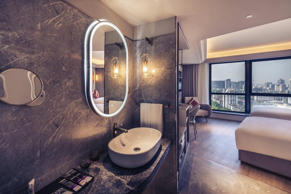 A modern hotel bathroom with a large oval mirror, a round sink, and a city view from the adjacent bedroom.