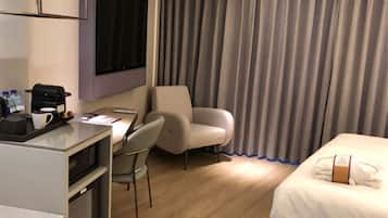 A hotel room with a bed, a desk with a coffee machine, a chair, and a TV mounted on the wall.