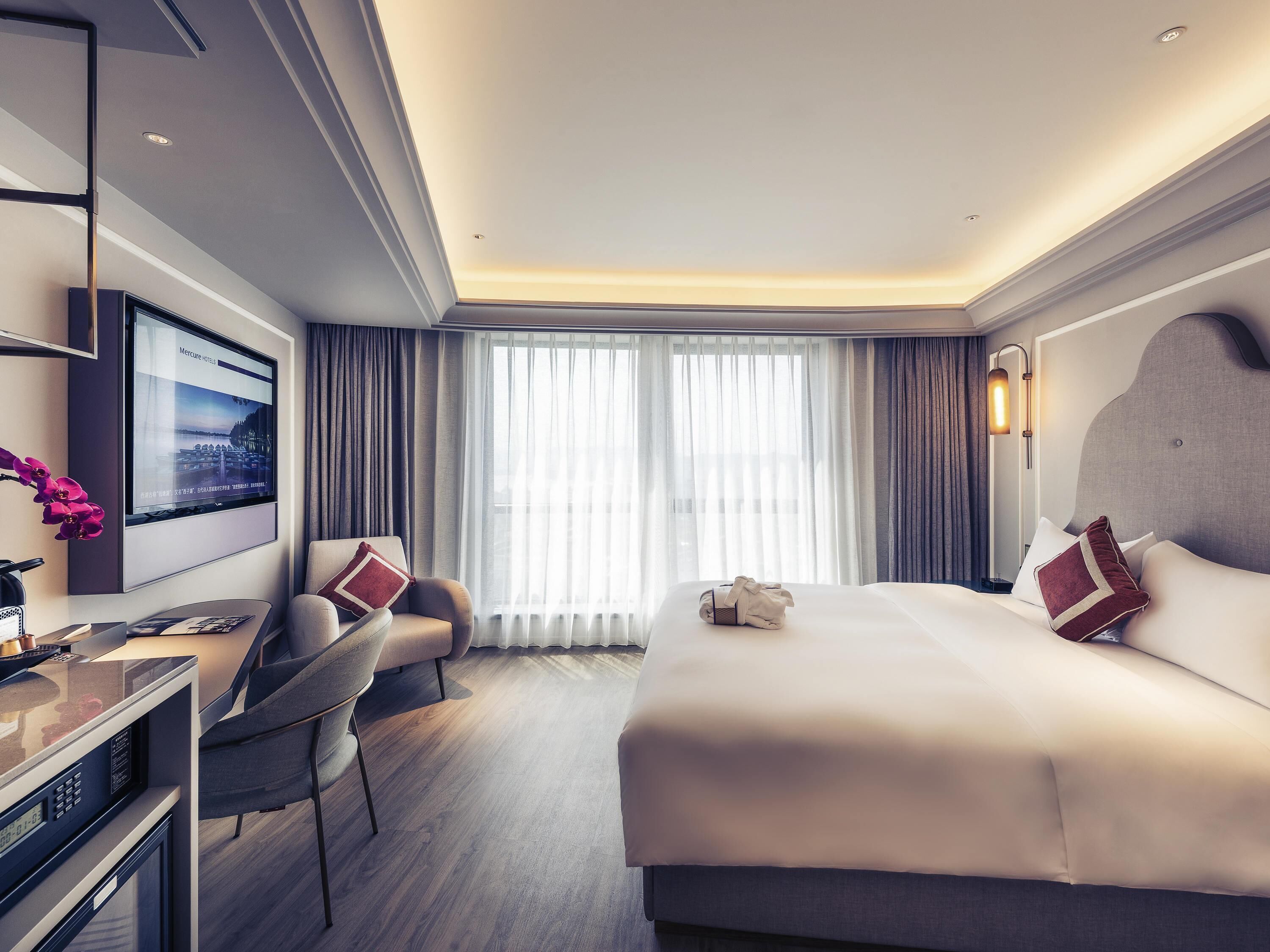 Une chambre d’hôtel moderne dotée d’un grand lit, d’un bureau, d’une chaise, d’une télévision et d’une grande fenêtre avec des rideaux.