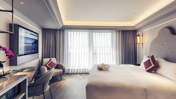 Una habitación de hotel moderna con una cama grande, un escritorio, una silla, un televisor y un ventanal con cortinas.