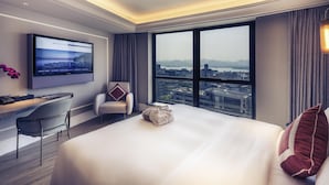 Une chambre d’hôtel avec un grand lit, un bureau avec une chaise, un téléviseur fixé au mur et une grande fenêtre donnant sur la ville.