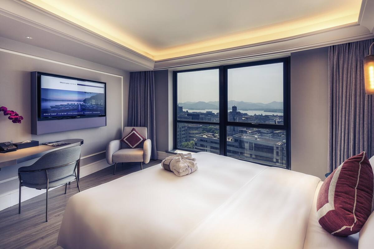 Une chambre d’hôtel avec un grand lit, un bureau avec une chaise, un téléviseur fixé au mur et une grande fenêtre donnant sur la ville.