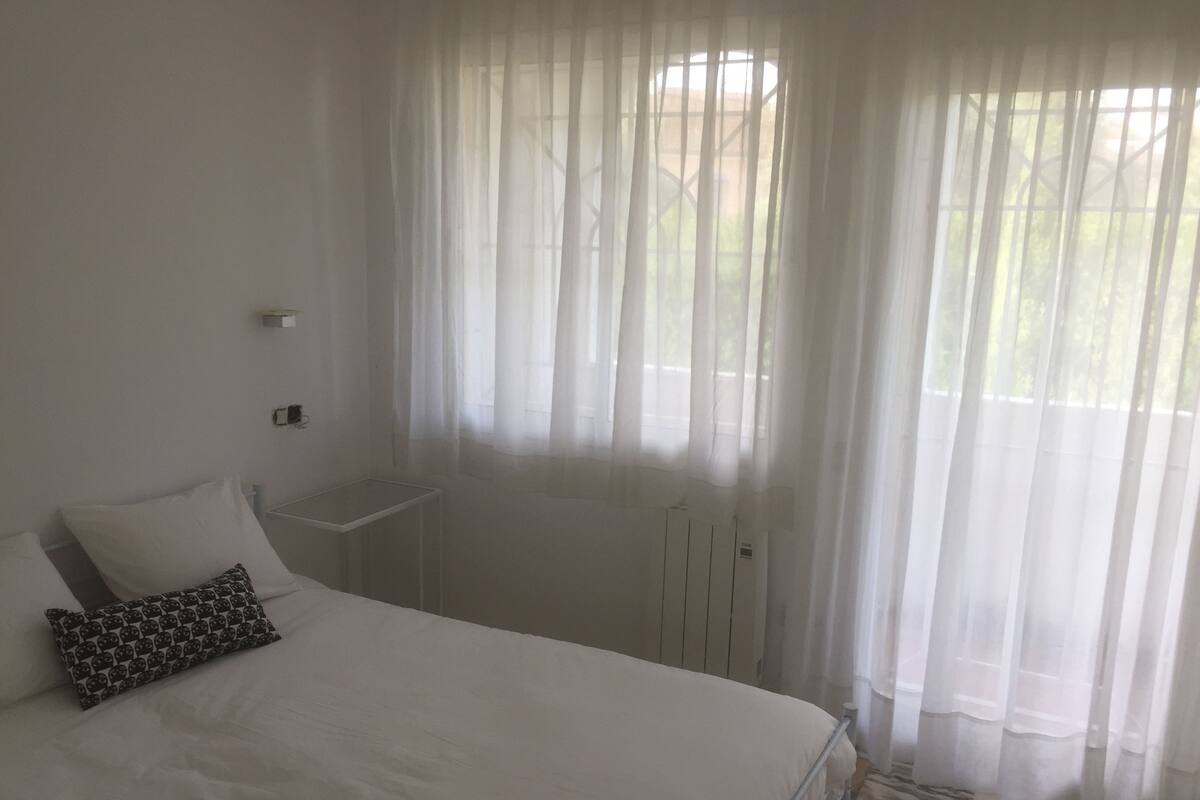 A bedroom with a bed, a bedside table, a lamp, and sheer curtains.
