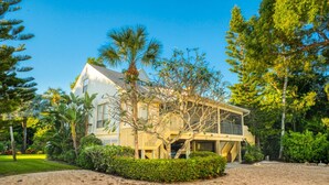 A two-story house with a sandy driveway, surrounded by lush greenery and tall trees.