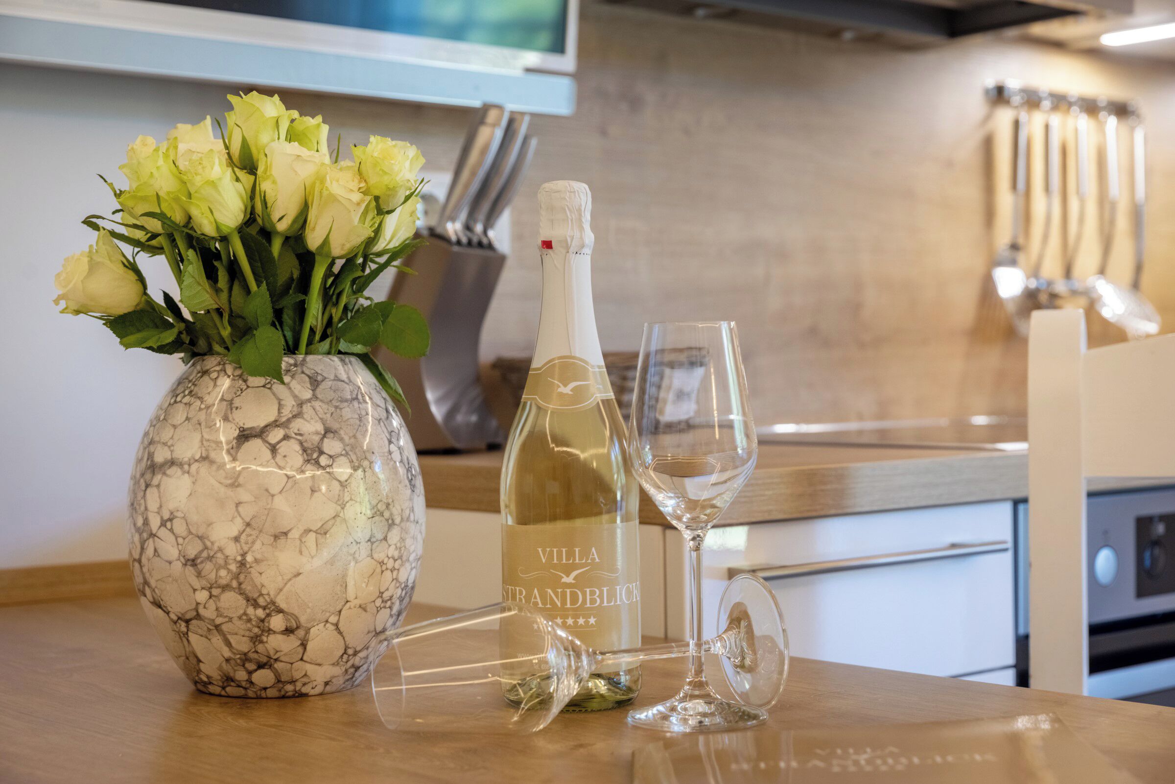Een marmeren vaas met gele rozen, een fles Villa Strandblick champagne en twee glazen op een aanrecht.