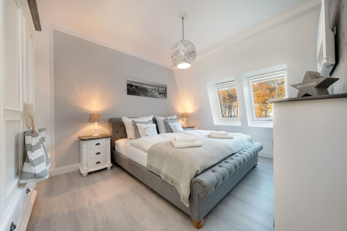 A bedroom with a grey upholstered bed, bedside tables with lamps, a framed picture on the wall, and a window with autumn foliage outside.