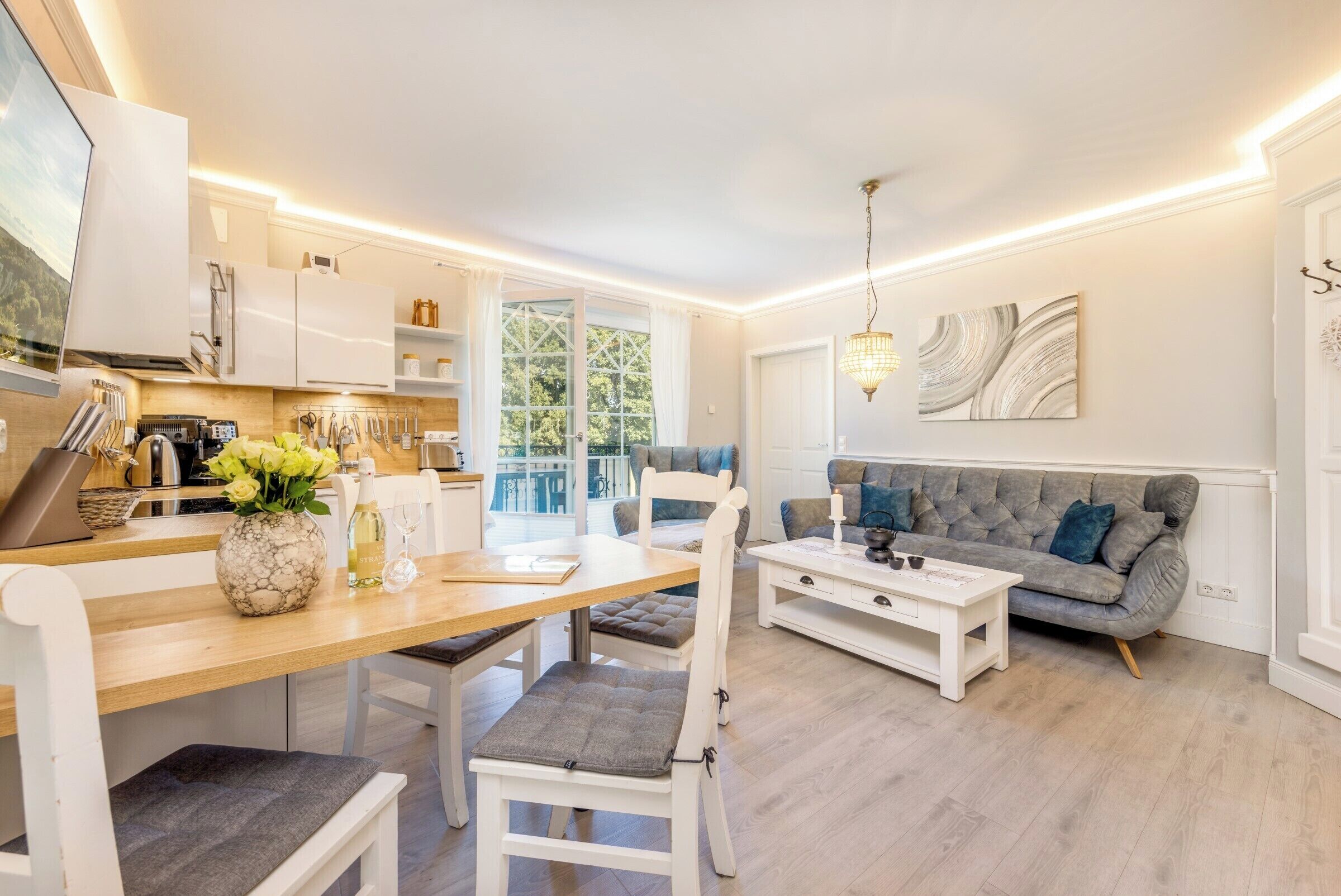 Une cuisine moderne avec des meubles blancs, un coin repas avec une table en bois et des chaises blanches, et un canapé gris dans un salon agrémenté d’un lustre.