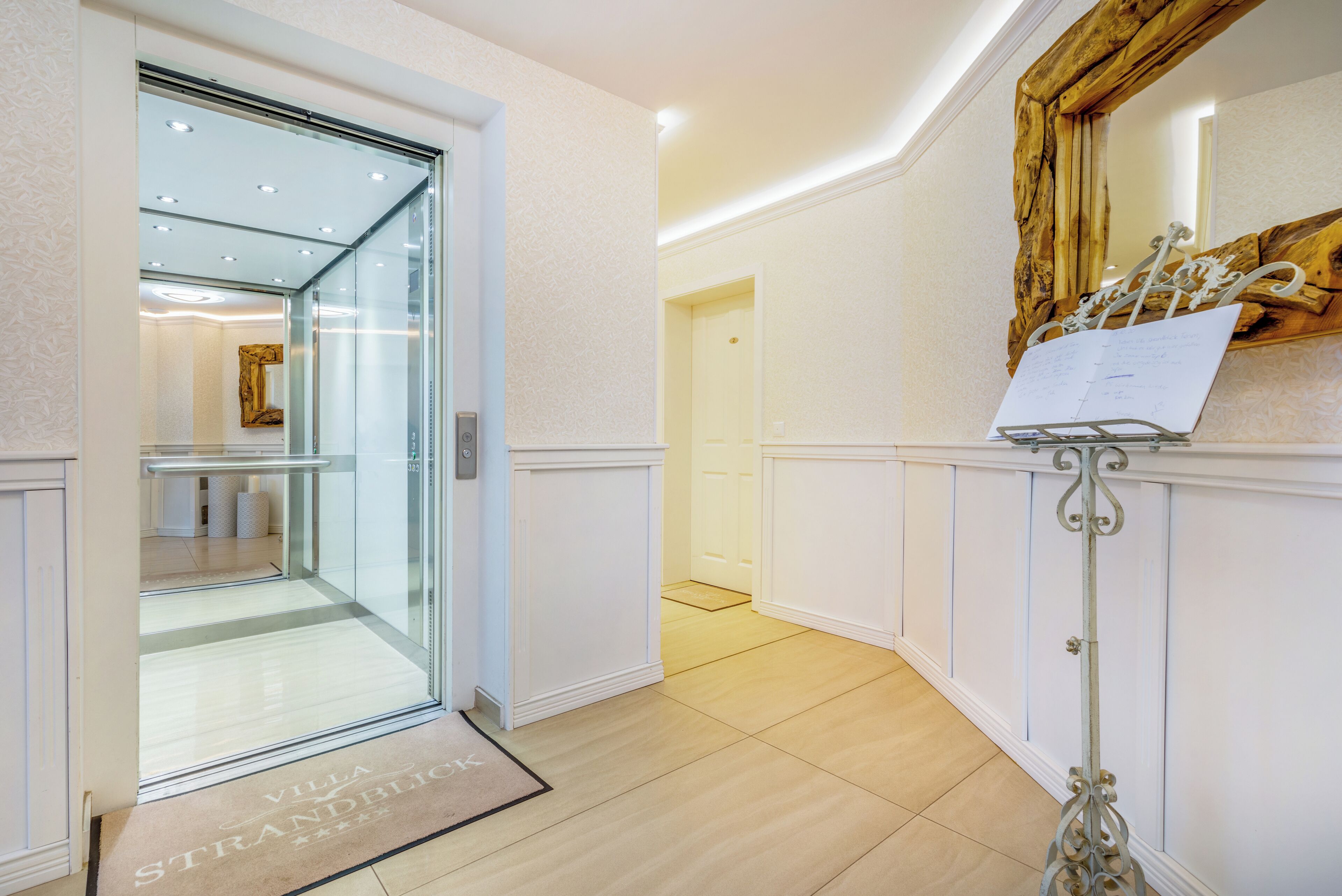 A hotel entrance with a glass door, a doormat, a metal stand with a paper, and a wooden-framed mirror on the wall.