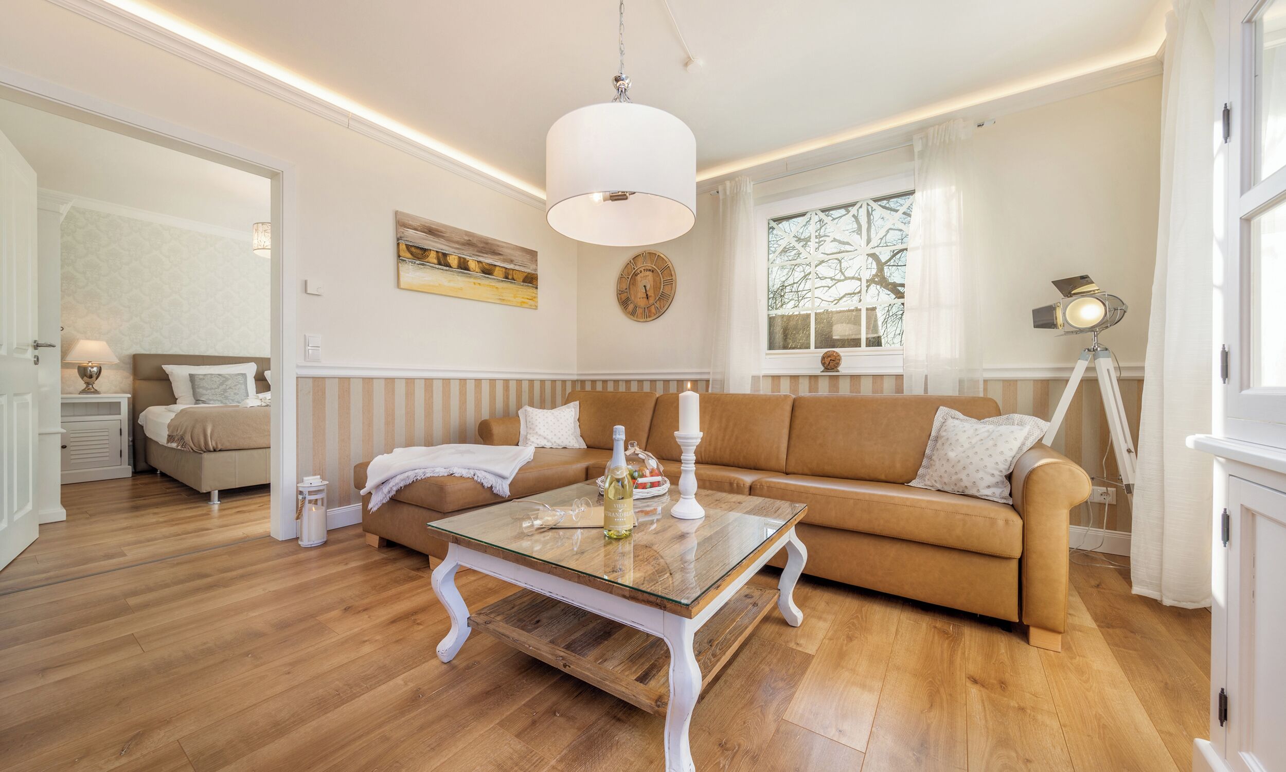 Un salón luminoso con un sofá marrón, una mesa de centro de cristal y una lámpara blanca.