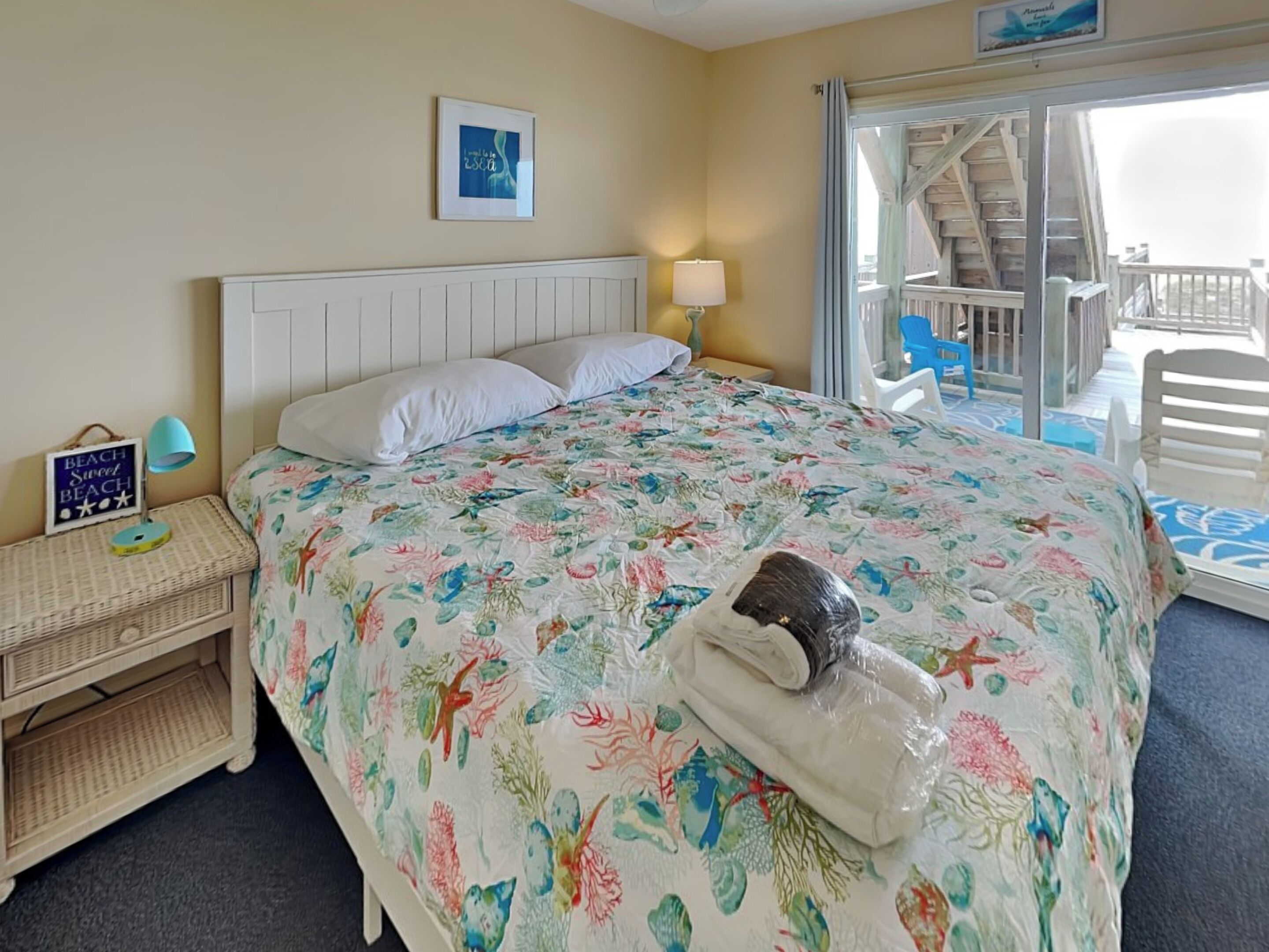 A bedroom with a bed, a nightstand, a lamp, a framed picture, and a view of a balcony with a chair.