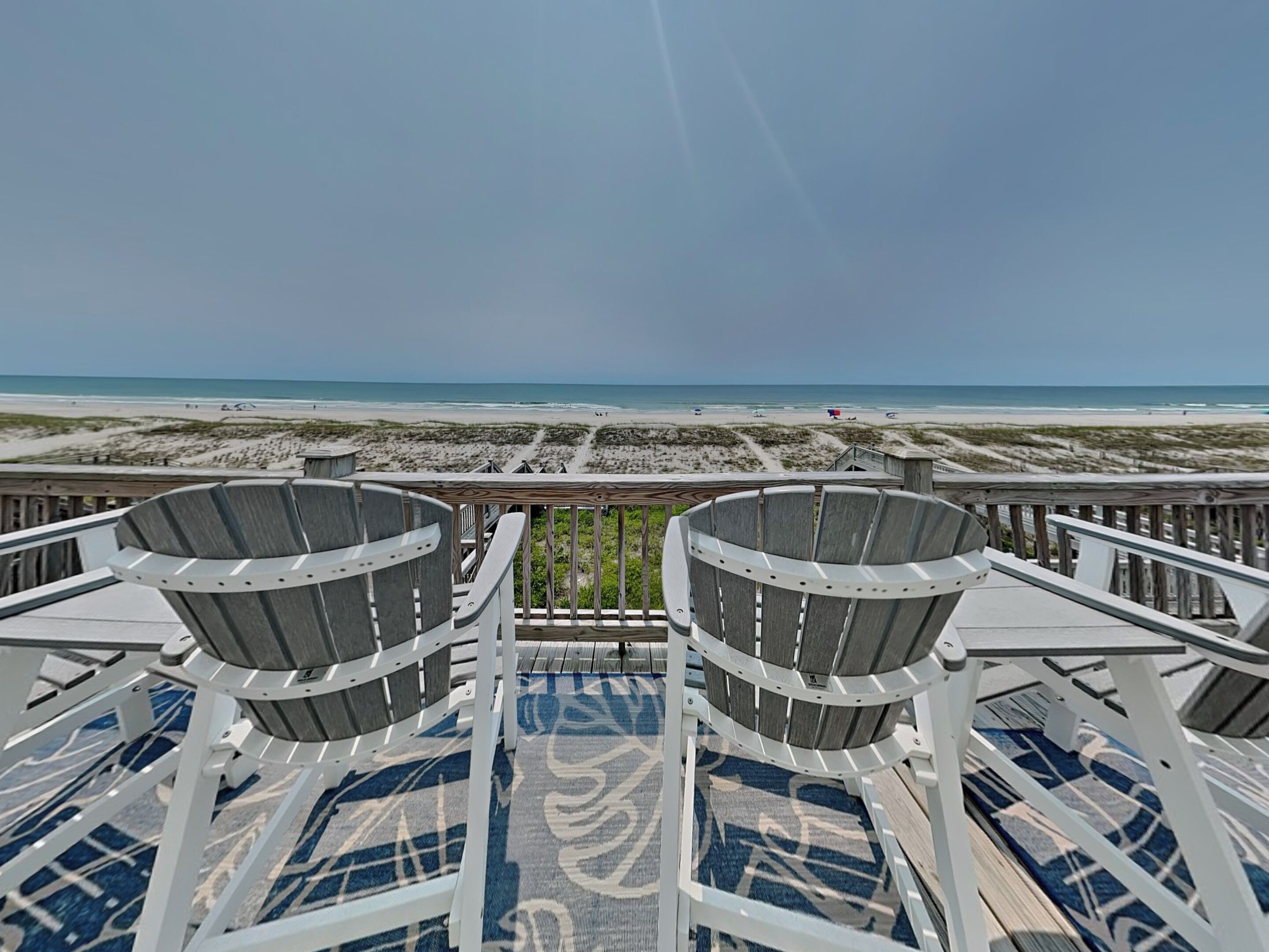 Une vue sur la plage depuis une terrasse équipée de deux chaises et d’un tapis à motifs.