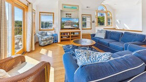 Ein geräumiges Wohnzimmer mit einem blauen Sofa, einem Korbstuhl und einem Couchtisch. Ein Fernseher ist an der Wand montiert und es gibt ein Fenster mit Blick ins Freie.