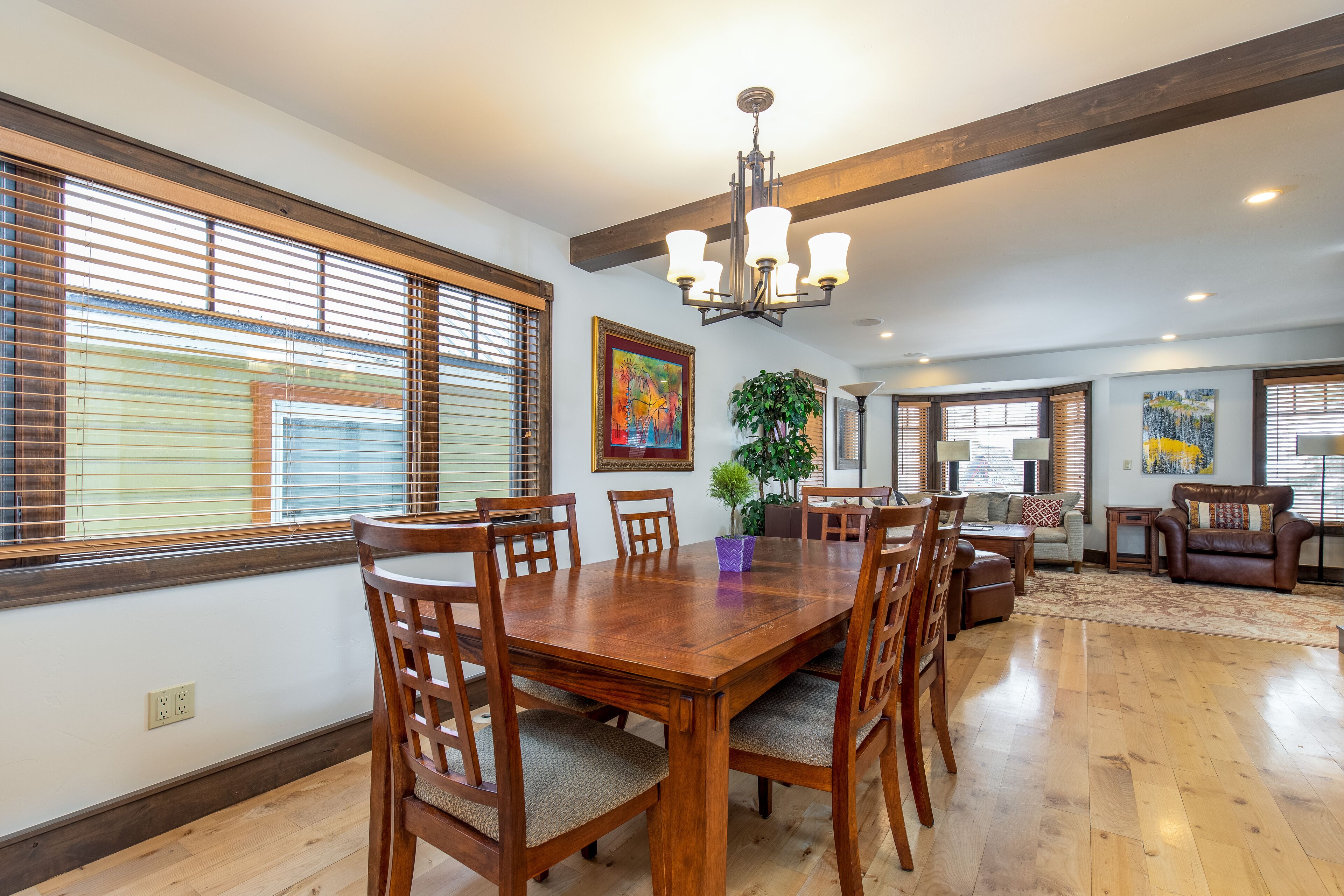 A spacious dining area with a wooden table and chairs, large windows with blinds, a chandelier, and artwork on the walls.