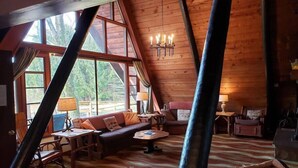 A cozy loft-style room with a large window, wooden beams, a sofa, a table, and a lamp. - Private Cabin with Lake & Mountain Views on 10 Acres - Near Resorts - Book Now! (Champion)