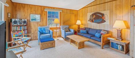 A cozy living room with wooden walls, a bookshelf, a blue armchair, a sofa, and two lamps.