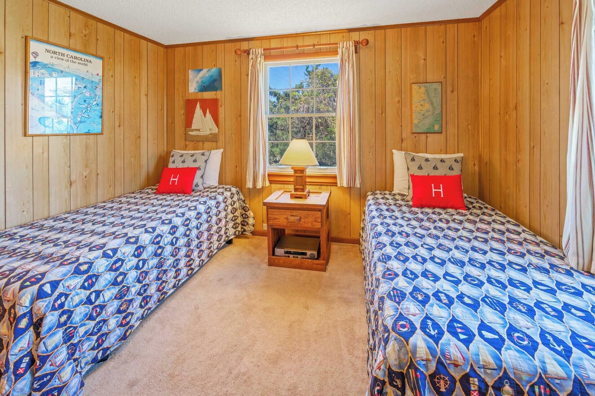 Una habitación con dos camas, cada una con una colcha azul y blanca de temática náutica y una almohada roja con la letra 'H'. En el centro, un mesita de noche de madera con una lámpara, una ventana con cortinas y paredes de madera.