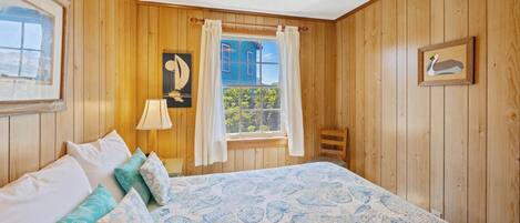 A wooden-paneled room with a bed, a window with curtains, a wall-mounted lamp, and a framed picture.