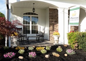 A building with a stone facade, a covered porch with chairs, and a flower bed with various flowers.