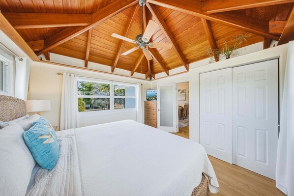 Un dormitorio con una cama grande, vigas de madera en el techo, un ventilador de techo y vistas del exterior a través de una ventana.