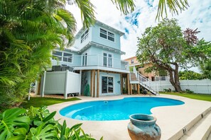 Una casa de dos pisos con piscina, rodeada de abundante vegetación y una cerca blanca.
