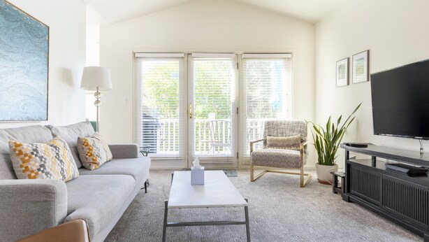 A living room with a grey sofa, a white coffee table, a black TV stand, and a large window with blinds. - Downtown Napa!  Unit C Walk to Everything!  (Napa)