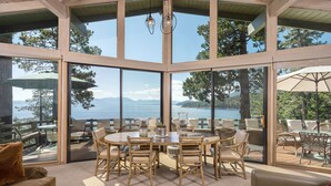 Un espacio interior amplio con una gran mesa redonda rodeada de sillas, ventanas grandes que ofrecen vistas a un lago y montañas, y un techo con vigas a la vista y luces colgantes.