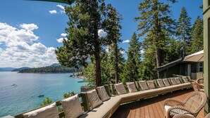 Terraza de madera con mobiliario exterior, con vista a un lago con barcos, rodeada de árboles altos.