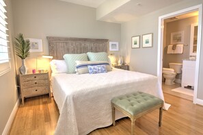 Une chambre à coucher avec un grand lit, des tables de chevet, une commode, un banc et une salle de bain visible à travers une porte ouverte.