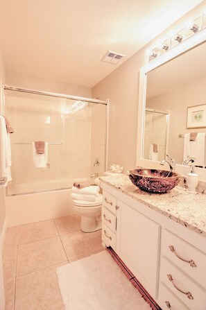 Une salle de bain dotée d’une douche à parois de verre, de toilettes blanches, d’un meuble-lavabo avec un grand miroir et d’un lavabo agrémenté d’un récipient décoratif.