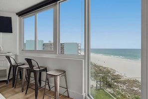 A room with a large window overlooking a beach, a desk with chairs, and a TV mounted on the wall.
