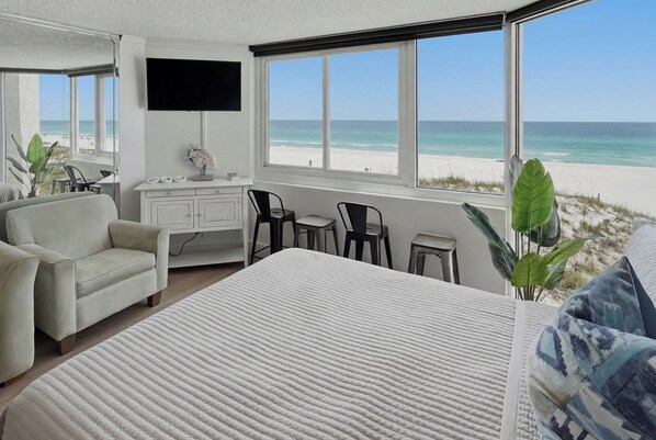 A bedroom with a bed, a desk, a TV, and a view of the beach.