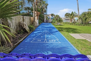 A blue shuffleboard court with numbers and an 'OFF' sign, surrounded by greenery and a wooden fence.