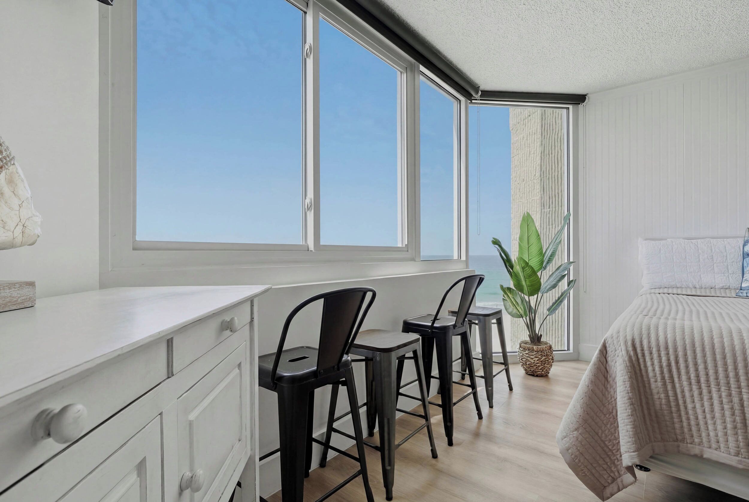 Une pièce avec une commode blanche, un lit recouvert d’une courtepointe, une grande fenêtre avec des rideaux et une plante en pot.