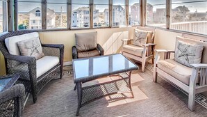 A sunlit room with wicker furniture, a glass coffee table, and large windows offering a view of residential buildings. - Semi-Oceanfront Home w/ Pool, Hot Tub, Elevator, Cabana w/ Tiki Bar, Game Room (Salvo)