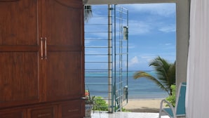 Een kamer met een houten garderobe, een bed met een bloemenprintdeken, uitzicht op een strand en zee door een glazen deur, en een palmboom.