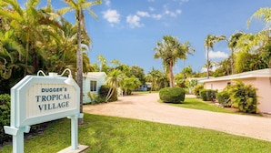 Ein Schild für Dugger's Tropical Village, mit einem Weg, der zu einer Reihe einstöckiger Gebäude inmitten von Palmen und üppiger Vegetation führt.