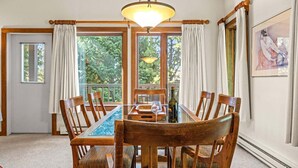 A dining area with a wooden table, chairs, a chandelier, and large windows with curtains. - RMR: Rendezvous A5 Condo in Teton Village (Teton Village)