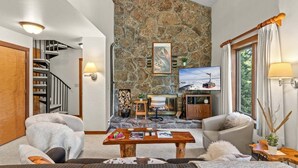 A cozy living room with a stone wall, a fireplace, a television, and a staircase. - RMR: Rendezvous A5 Condo in Teton Village (Teton Village)