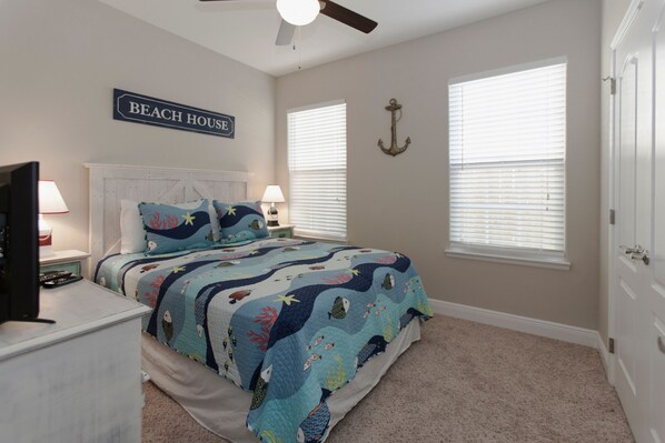A bedroom with a bed, a nightstand, a television, a ceiling fan, and a sign that reads "BEACH HOUSE".