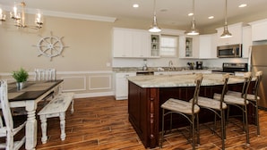A kitchen with wooden flooring, white cabinetry, a central island with bar stools, pendant lighting, and a ship's wheel decoration. - Walk just 2 blocks to the beach! (PANAMA CITY BEACH)