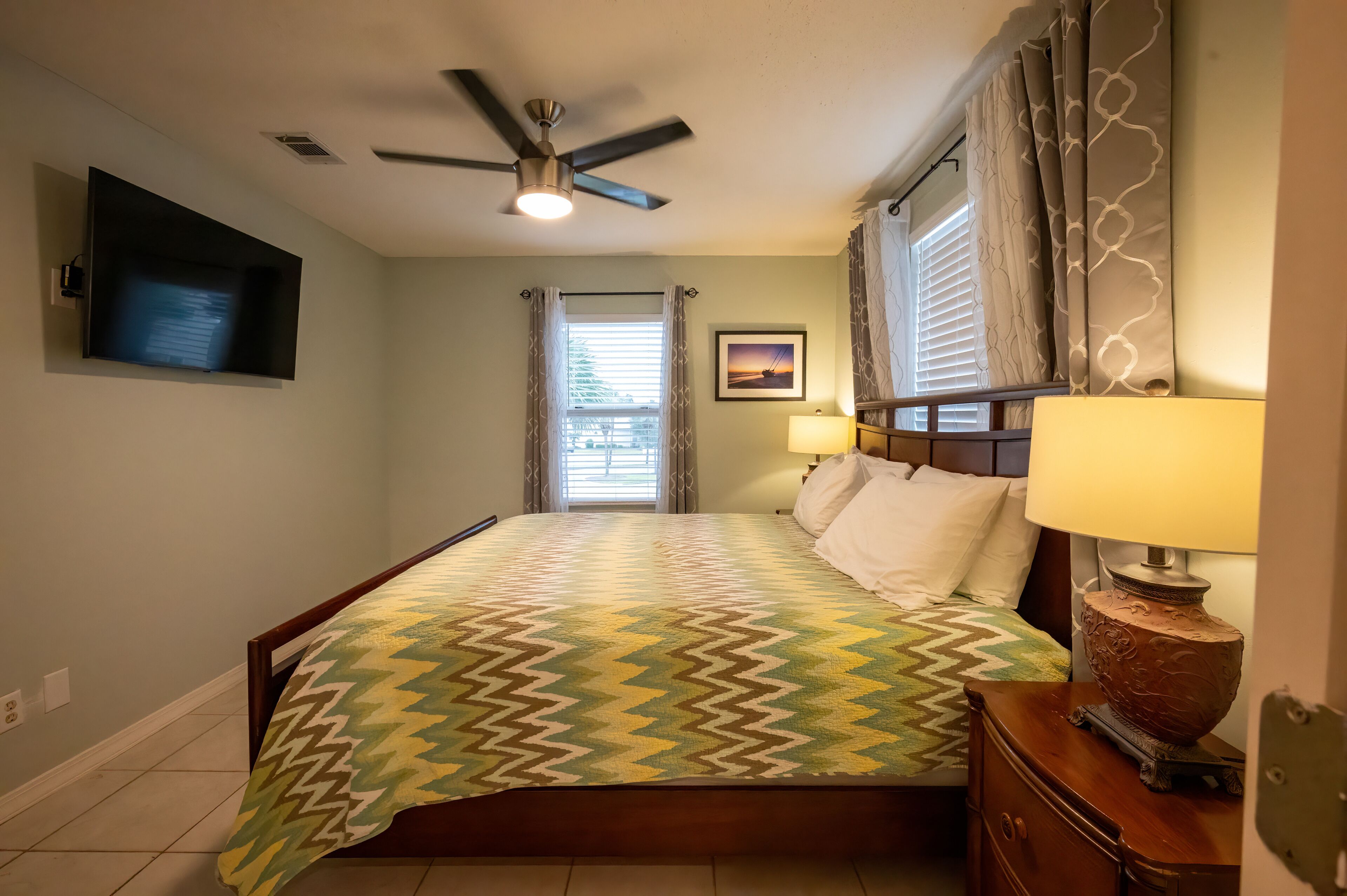 A bedroom with a bed, a ceiling fan, a TV mounted on the wall, a window with curtains, and a bedside lamp.
