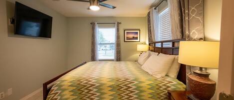 A bedroom with a bed, a ceiling fan, a TV mounted on the wall, a window with curtains, and a bedside lamp.