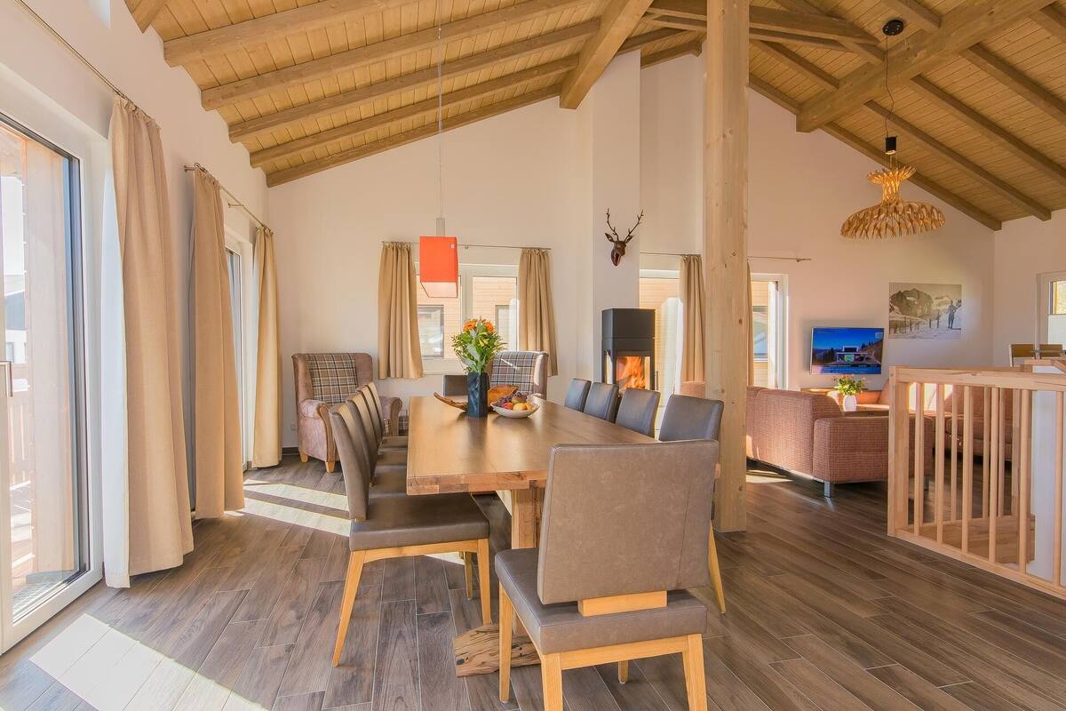 Een ruime woonkamer met hoog houten plafond, een open haard en een eetgedeelte met een lange tafel en stoelen.