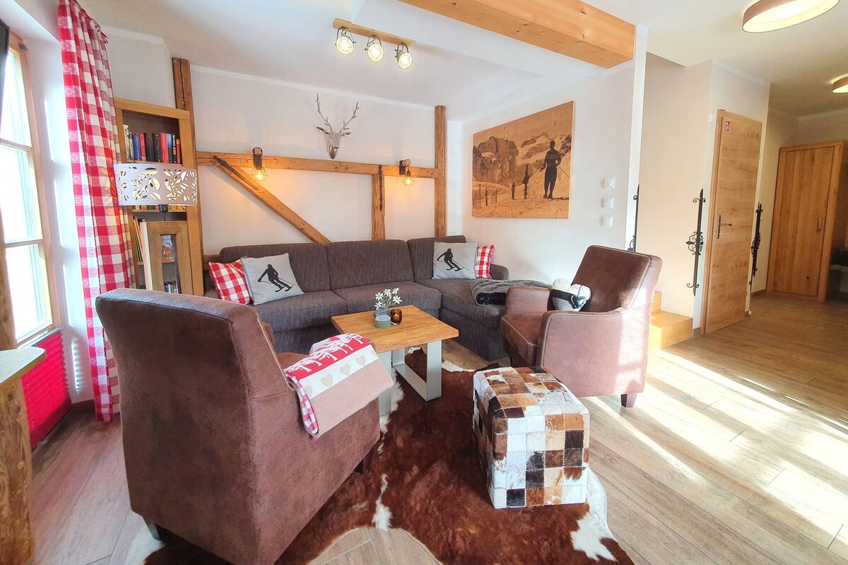 Een gezellige woonkamer met een bank, twee fauteuils, een salontafel en een decoratief hertenhoofd. Er is een houten plafond, een schilderij en een boekenplank vol boeken.