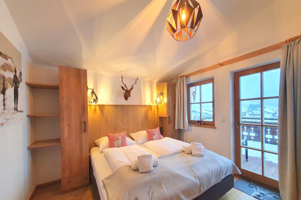 Een slaapkamer met een groot bed, een houten hoofdbord en een hertenhoofd aan de muur. Er is een raam met gordijnen en uitzicht op een besneeerd landschap.