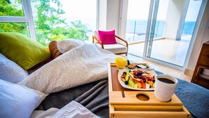 Une chambre avec un lit, un plateau de petit-déjeuner avec des fruits, une tasse de café et une vue sur la mer par de grandes fenêtres.